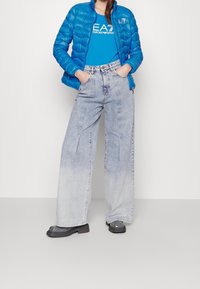 Niebieska kurtka puchowa o błyszczącym wykończeniu, zestawiona z jasnoniebieskimi, szerokimi spodniami jeansowymi. Czarne sandały na platformie o gładkiej powierzchni.