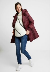 Μπουφάν μπουφάν μπορντό με κουκούλα, φορεμένο πάνω από πουλόβερ σε κρεμ απόχρωση με διαγώνιο μαύρο μοτίβο, σε συνδυασμό με μπλε τζιν και λευκά αθλητικά παπούτσια.