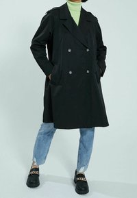 Czarny dwurzędowy trencz z dużymi guzikami, kieszeniami po bokach i epoletami na ramionach; zestawiony z przetartymi, jasnoniebieskimi dżinsami i czarnymi butami.