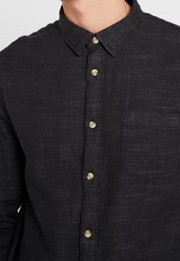 Musta napillinen paita, joka on valmistettu teksturoidusta kankaasta. Paidassa on kaulus, vaaleat napit ja yksi rintatasku.