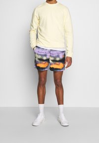 Żółta bluza z okrągłym dekoltem, spodenki tie-dye w wielokolorowe wzory z fioletowym, pomarańczowym i czarnym, w połączeniu z białymi trampkami i skarpetkami.