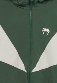 Πράσινο μπουφάν με λευκές λεπτομέρειες, που διαθέτει φερμουάρ ολόκληρου μήκους και λογότυπο. Λείο ύφασμα με γεωμετρικά σχέδια στο μπροστινό μέρος.