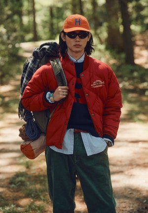 Jeune personne portant une casquette orange et une veste rouge, portant un sac à dos et du matériel de camping sur un sentier forestier.