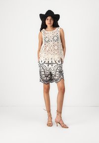 Top in cotone all'uncinetto di colore bianco sporco con fori floreali, abbinato a pantaloni corti ricamati in bianco e nero con orlo a scallop. Tacchi marroni con strappi.