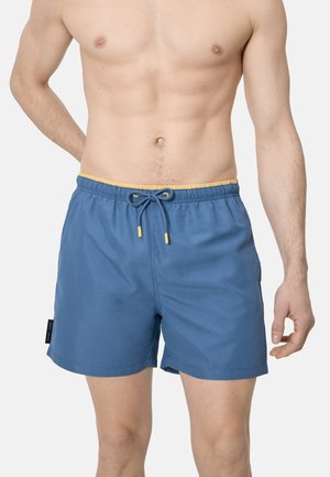 Torso masculino con pantalones cortos de baño azules con cintura y cordón amarillos, de pie frente a un fondo liso.