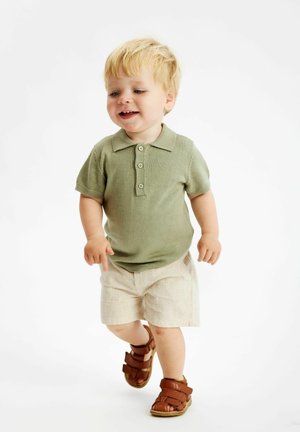 Ljusgrön stickad polo med tre knappar, ihop med beige shorts och bruna lädersandaler, på ett gående barn. Enkel bakgrund.