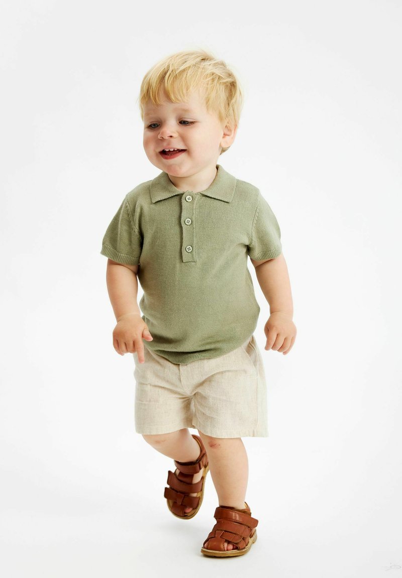 Ljusgrön stickad polo med tre knappar, ihop med beige shorts och bruna lädersandaler, på ett gående barn. Enkel bakgrund.