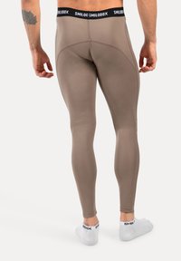 Man in taupe fitted leggings en witte enkelsokken met zwart "SMILODOX" merk, staand op blote voeten op een witte vloer.