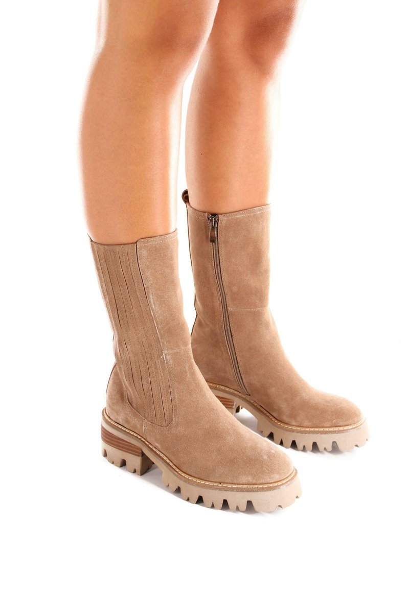 Botas de ante color arena hasta la rodilla con suela de goma gruesa, que cuentan con una cremallera lateral y acentos verticales en relieve a lo largo del cañón.