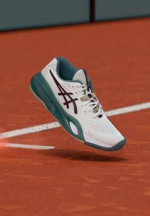 Hvid og grøn tennissko i luften over en rød lerbane, der puffer lerpartikler op nær hvide linjemarkeringer.