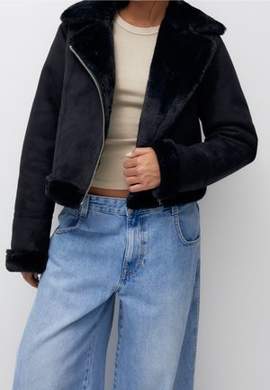 Femme portant une veste noire doublée fausse fourrure, un crop top beige et un jean ample bleu clair sur fond uni.