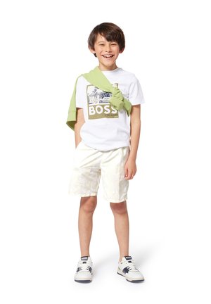 Weißes T-Shirt mit grünem "BOSS"-Grafik, hellgelbe gemusterte Shorts und grüner Pullover über die Schultern gelegt. Weiße und marineblaue Sneakers.