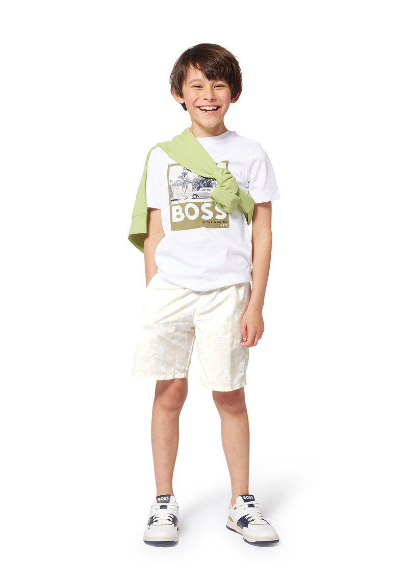 T-shirt blanc avec un motif vert "BOSS", short à motifs jaune clair, et un pull vert drapé sur les épaules. Baskets blanches et navy.