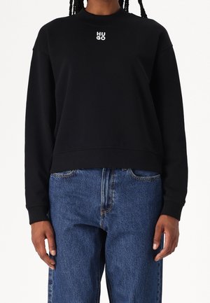 Personne portant un sweat-shirt noir à manches longues avec un texte blanc "HUGO" et un jean bleu, debout devant un fond blanc uni.