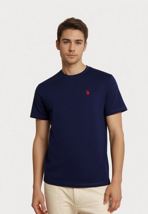 Junger Mann trägt ein marineblaues T-Shirt mit Rundhalsausschnitt und einem kleinen rot gestickten Logo auf der linken Brust, kombiniert mit beigen Hosen.