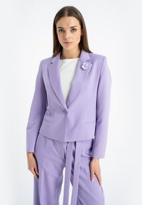Liliowy żakiet o dopasowanym kroju, zapinany na jeden guzik, z dwiema przednimi kieszeniami, ozdobiony kwiatową broszką na klapie.