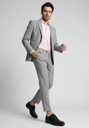 Mand i lyst gråt jakkesæt med lyserød skjorte går selvsikkert, iført sorte festlige sko mod ensfarvet grå baggrund.