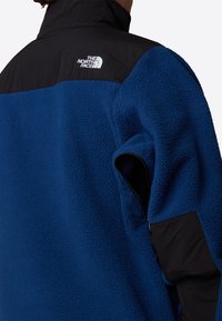 Niebieska polarowa kurtka z czarną nylonową górną częścią. Wśród cech znajdują się wysoki kołnierz, kieszeń na zamek oraz logo marki na ramieniu.