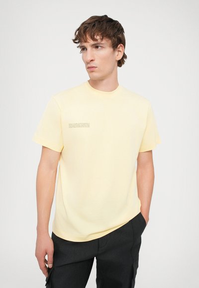 Tricou din bumbac galben, cu guler rotund și mâneci scurte. Include detalii imprimate pe partea stângă. Poartă cu pantaloni închiși la culoare.