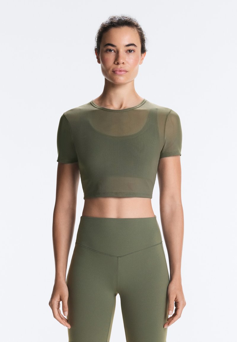 Jeune femme de face portant un crop top en mesh vert olive et des leggings taille haute assortis sur fond blanc uni.