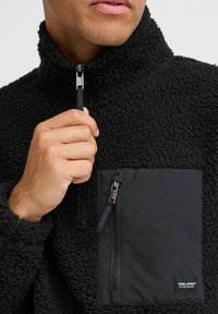 Black fleece pullover with a high collar, featuring a zippered chest pocket made of smooth fabric. Zipper detail on the collar and pocket.