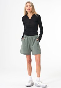 Schwarzes Langarmshirt mit Reißverschluss und grünen Shorts mit elastischem Bund, die seitliche Taschen und ein schlichtes Design aufweisen. Weiße Sneakers.