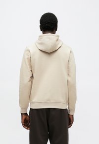 Rövid göndör hajú férfi bézs kapucnis pulóverben és sötét nadrágban, egy sima fehér háttér előtt állva, szemben a nézővel.