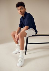 Granatowa koszulka polo z krótkim rękawem, jasnoszortkie szorty, białe trampki i białe skarpetki. Siedzi na czarnym krześle na beżowym tle.