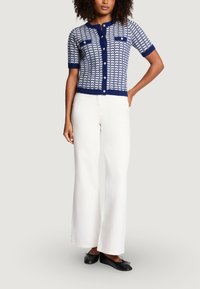 Granatowy i biały wzorzysty sweter na drutach z krótkimi rękawami, z dwoma przednimi kieszeniami i białymi guzikami, w połączeniu z białymi spodniami o szerokich nogawkach.