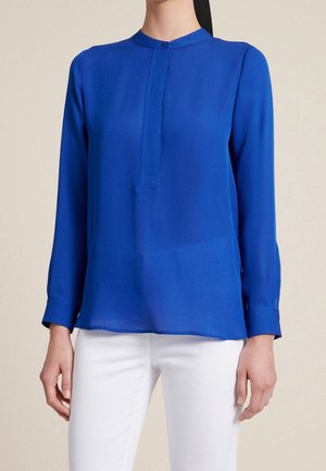 Mujer que lleva una blusa de manga larga azul real con un pequeño cuello redondo y pantalones blancos contra un fondo neutro.
