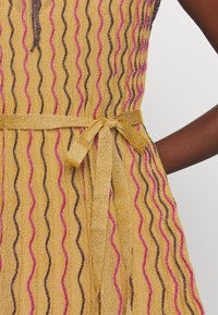 Close-up van een mosterdgele jurk met roze en zwarte golvende strepen, voorzien van een gestrikte riem in de taille en een donkere huidarm die op de heup rust.