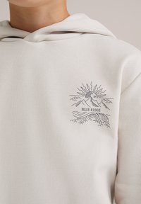 Crèmekleurige hoodie met een zwarte lijntekening van een zon, bergen en golven, waarbij de tekst "BLUE RIDGE" in het ontwerp is geïntegreerd.