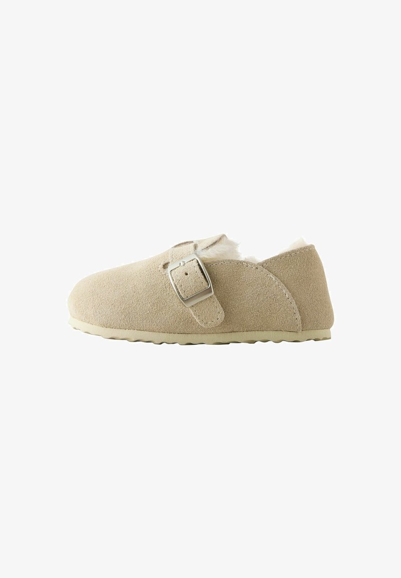 Chaussure beige en daim à enfiler avec une bride à boucle argentée et une doublure blanche duveteuse, vue de côté sur fond blanc.