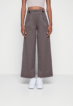 Höga, vida byxor i brun houndstooth-mönster, med två framsittande knappar och sidofickor. Vita sneakers fulländar looken.