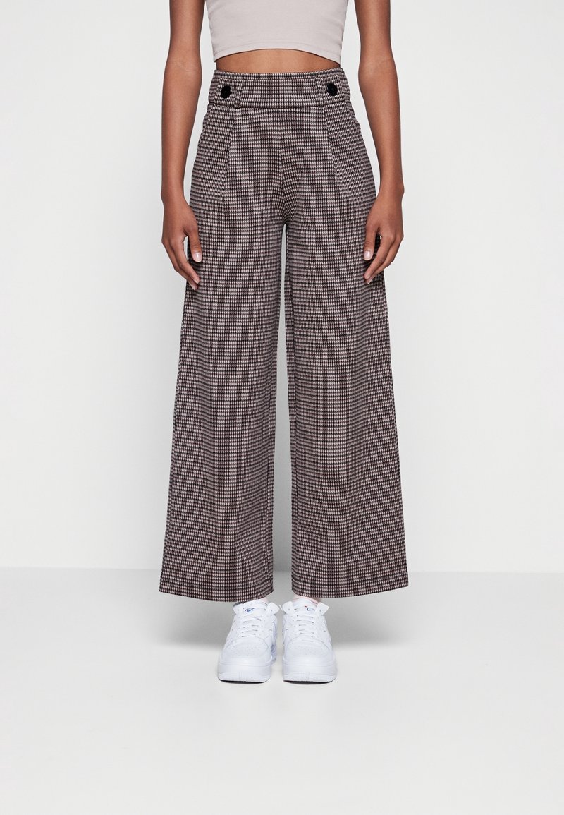 Höga, vida byxor i brun houndstooth-mönster, med två framsittande knappar och sidofickor. Vita sneakers fulländar looken.