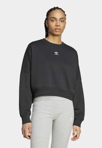 Černá cropped mikina s dlouhými rukávy a ribbovaným lemem, s bílým logem Adidas na hrudi. Nosit s přiléhavými šedými legínami.