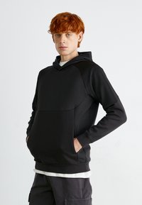 Felpa nera realizzata in un tessuto liscio e elasticizzato. Presenta una tasca a marsupio, un cappuccio regolabile con cordino e un design aderente.