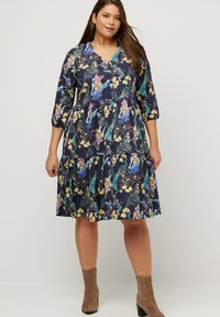 Abito a stampa floreale in tessuto blu scuro, con maniche a tre quarti, gonna a strati e scollatura a V. Indossato con stivali alla caviglia beige.