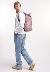 Plecak z rolowanym topem w miękkim różowym materiale, prostokątny kształt z wyściełanymi paskami, prezentowany na osobie ubranej w białą koszulę z długim rękawem oraz niebieskie dżinsy.