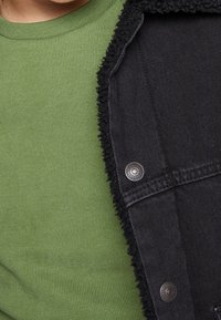 Černá džínová bunda s texturovaným límcem, se stříbrnými patentkami a pod ní zelené bavlněné tričko.