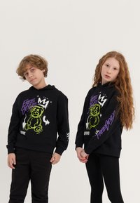 Sweatshirts pretos com capuz, apresentando gráficos de ursos de pelúcia em amarelo neon e texto roxo. Tecido macio com um corte descontraído, ideal para uso casual.