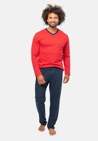 Rode langemouwen shirt met een V-hals en marineblauwe accenten, gecombineerd met marineblauwe lichtgewicht broeken, tentoongesteld op een model dat blootsvoets staat.