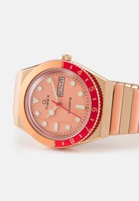 Montre Timex à quartz avec un boîtier en or rose, un cadran rose, une lunette rouge, des index horaires blancs et un bracelet en métal avec une finition brossée.