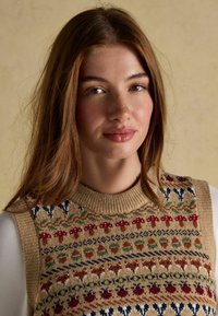Bež soe kootud ilma varrukateta sviiter mitmevärviliste mustritega, sealhulgas lilled ja südamed, ning ribitud ümmargune kaelus. Pehme tekstuur.