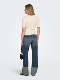 Žena okrenuta leđima, odjevena u kremasti top s kratkim rukavima, plave traperice sa širokim sivim manžetnama, crne cipele i drži crnu torbu.