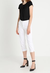 Kobieta w czarnej bluzce z krótkim rękawem, białych trapezowych spodniach z przednimi guzikami i czarnych wysokich sandałach na pasku wokół kostki stoi.