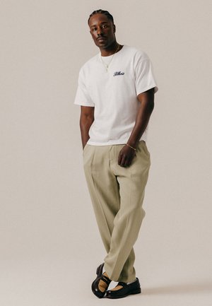 Homme aux cheveux tressés portant un t-shirt blanc, un pantalon beige, des chaussures noires et des bijoux en or, debout les mains dans les poches.