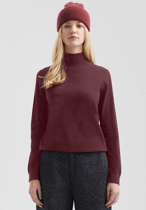 Femme portant un pull à col roulé en tricot bordeaux, un bonnet assorti et un pantalon foncé à motifs, debout devant un fond gris clair.