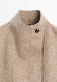 Beige wool coat with a high collar and a single visible dark button near the neck; minimal seam detailing on a plain background.