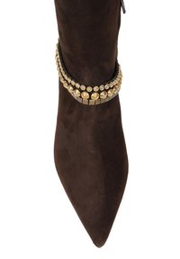 Brązowy zamszowy botek o spiczastym nosku z metalowymi zdobieniami wokół kostki, z mieszanką złotych akcentów i okrągłych wzorów.